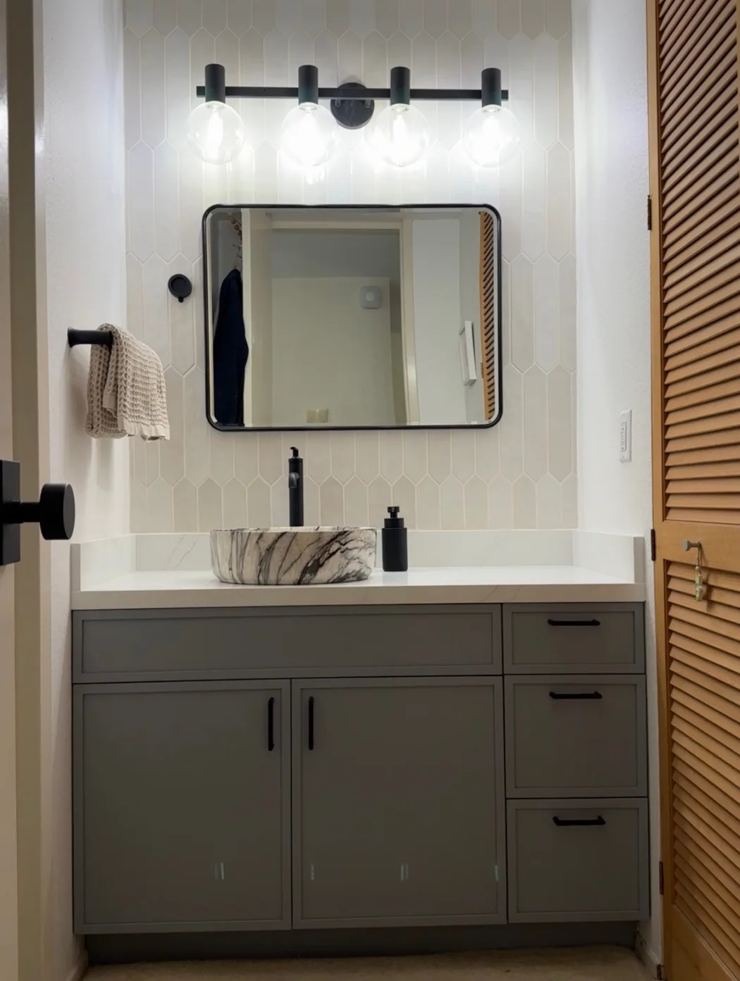 Bathroom vanity with drawers, a stone vessel basin, a black framed mirror and a four globe light, beside a louvered door