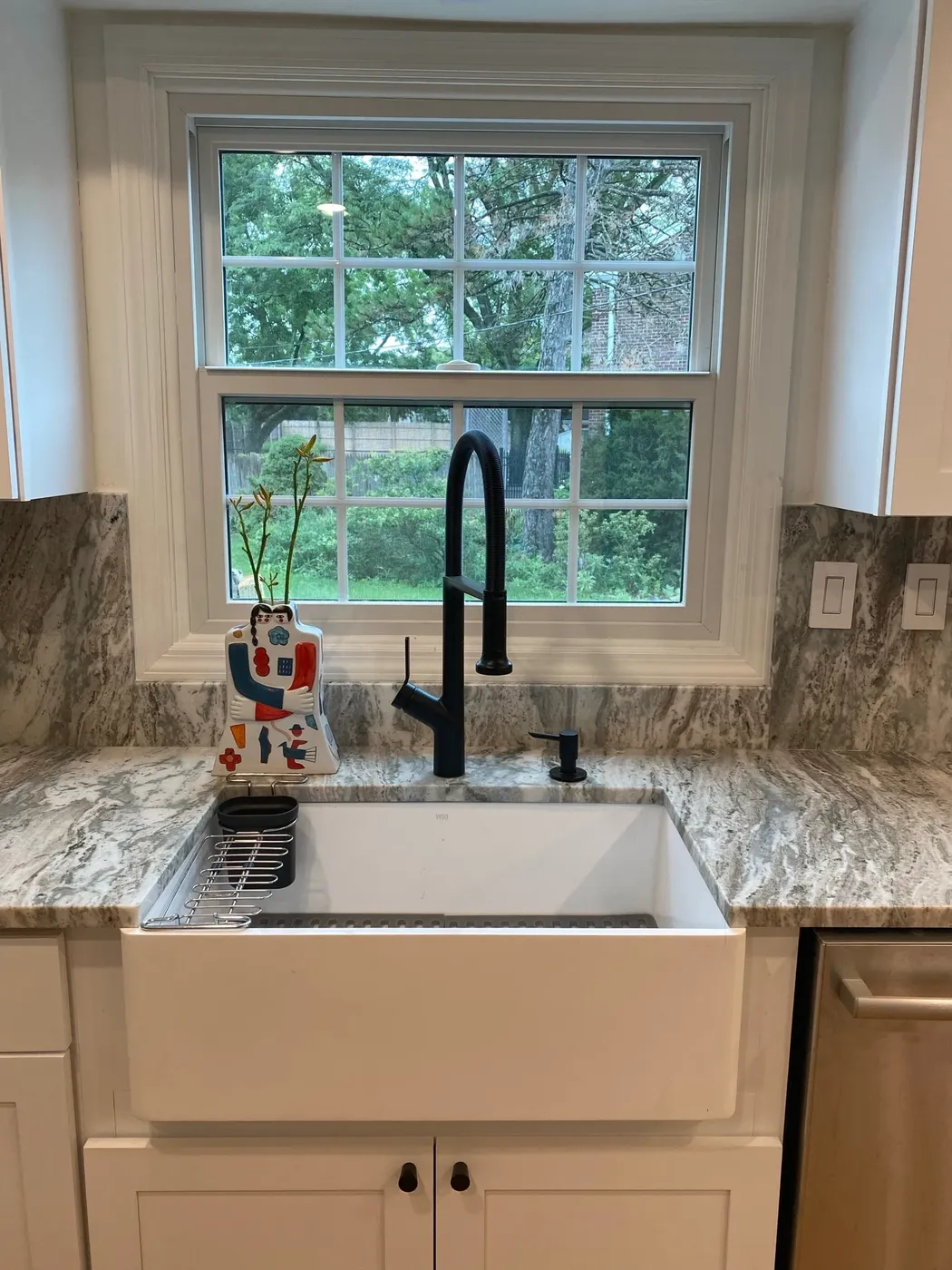 Apron-front farmhouse sink under a window with a matte black gooseneck tap and stone counter